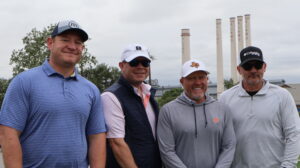 Four men posing outdoors near industrial smokestacks