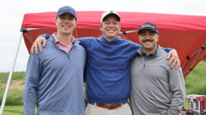 Three men smiling under red canopy at golf course