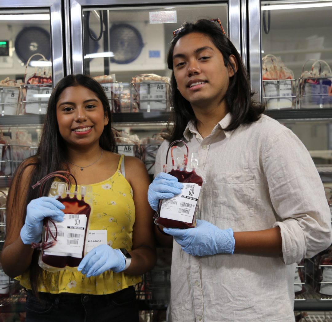 Olivia and Jacob, sickle cell fighters, hold up blood bags in the refrigerated inventory area.